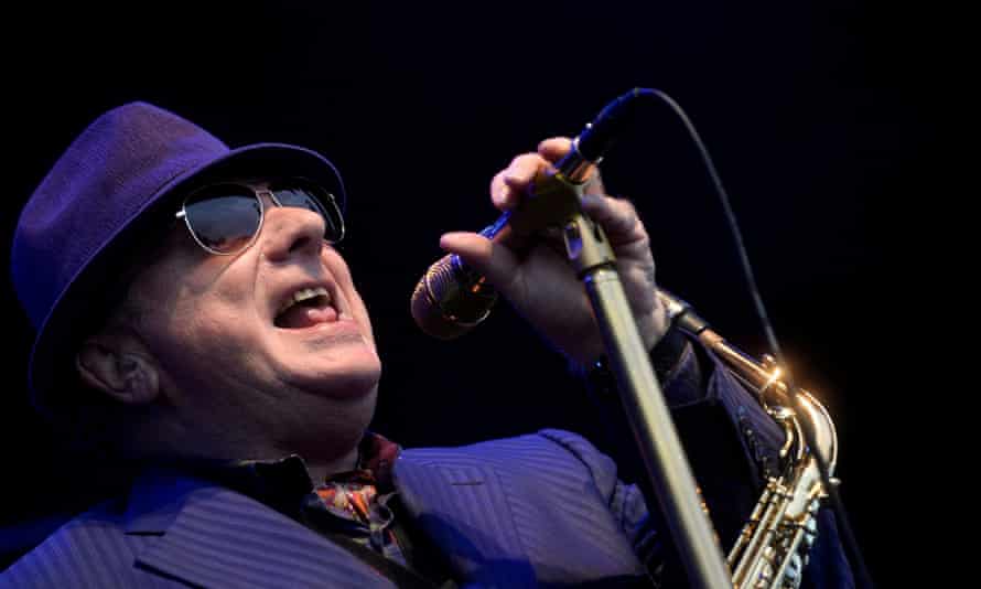Singer-songwriter Van Morrison wearing sunglasses, a deep purple suit and matching hat, holding a microphone to his mouth as he tilts his head back.