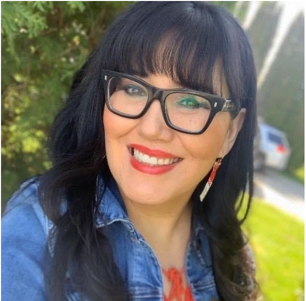 ID: Annita smiles with eyeglasses, beaded earring and denim jacket near green forest